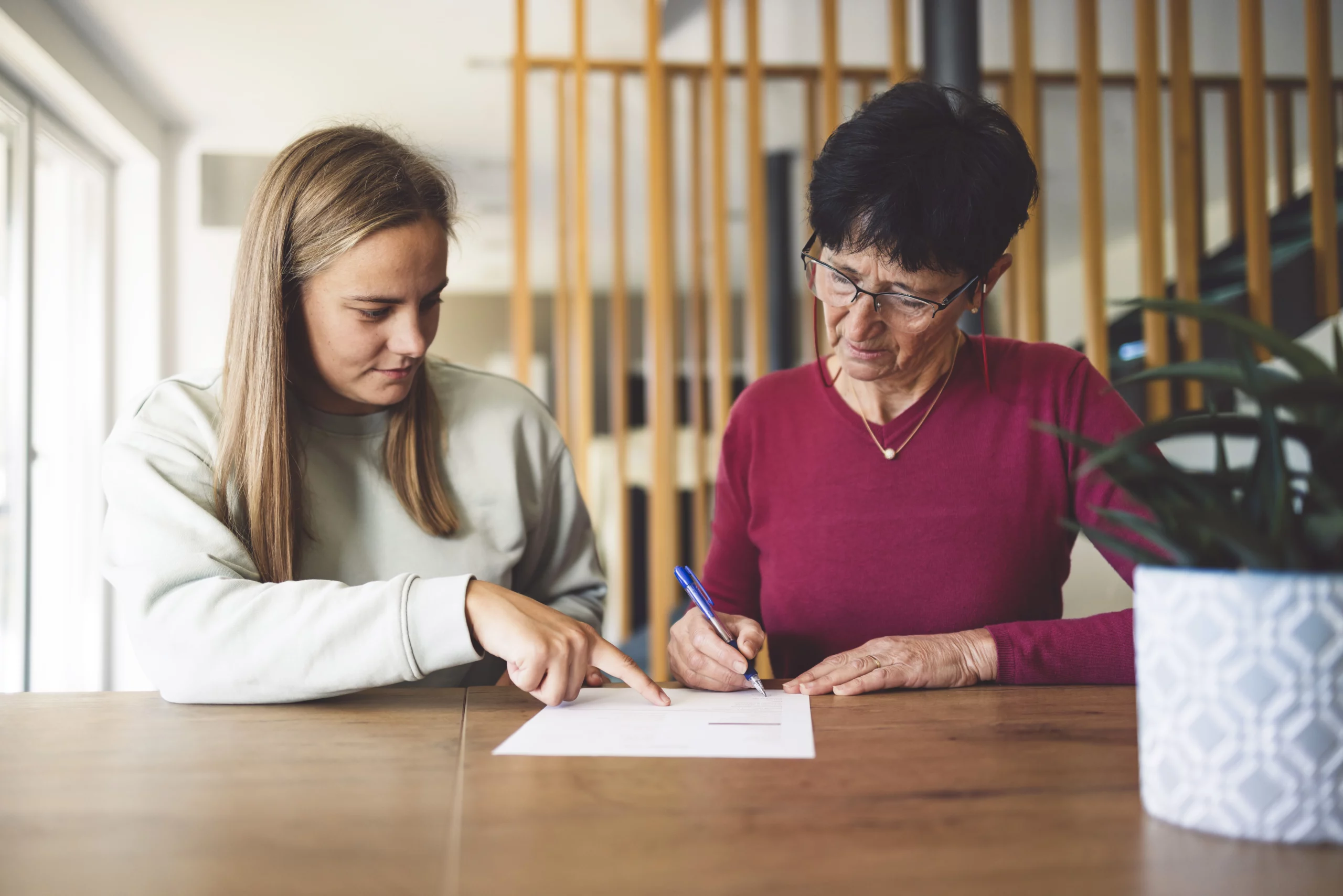 Young woman assisting an older woman with signing legal documents at a table, highlighting potential undue influence concerns.
