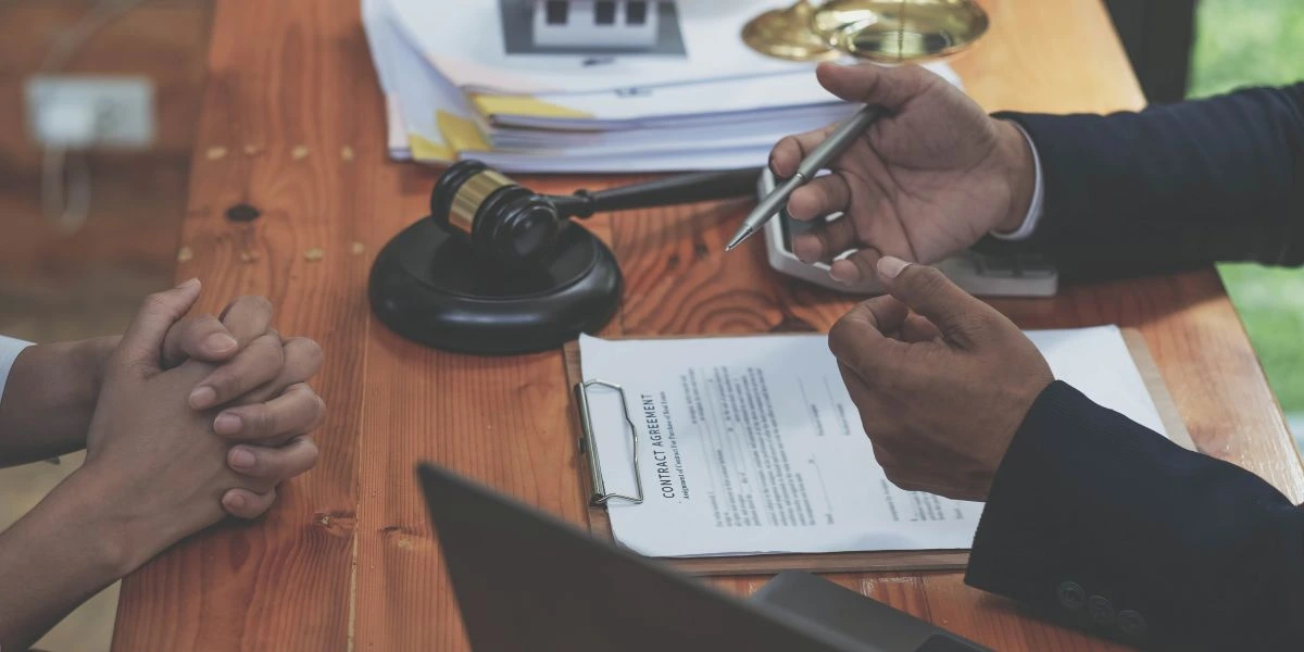Close-up of a lawyer discussing a contract agreement with a client, showing legal documents, gavel, and scales of justice on a wooden desk.
