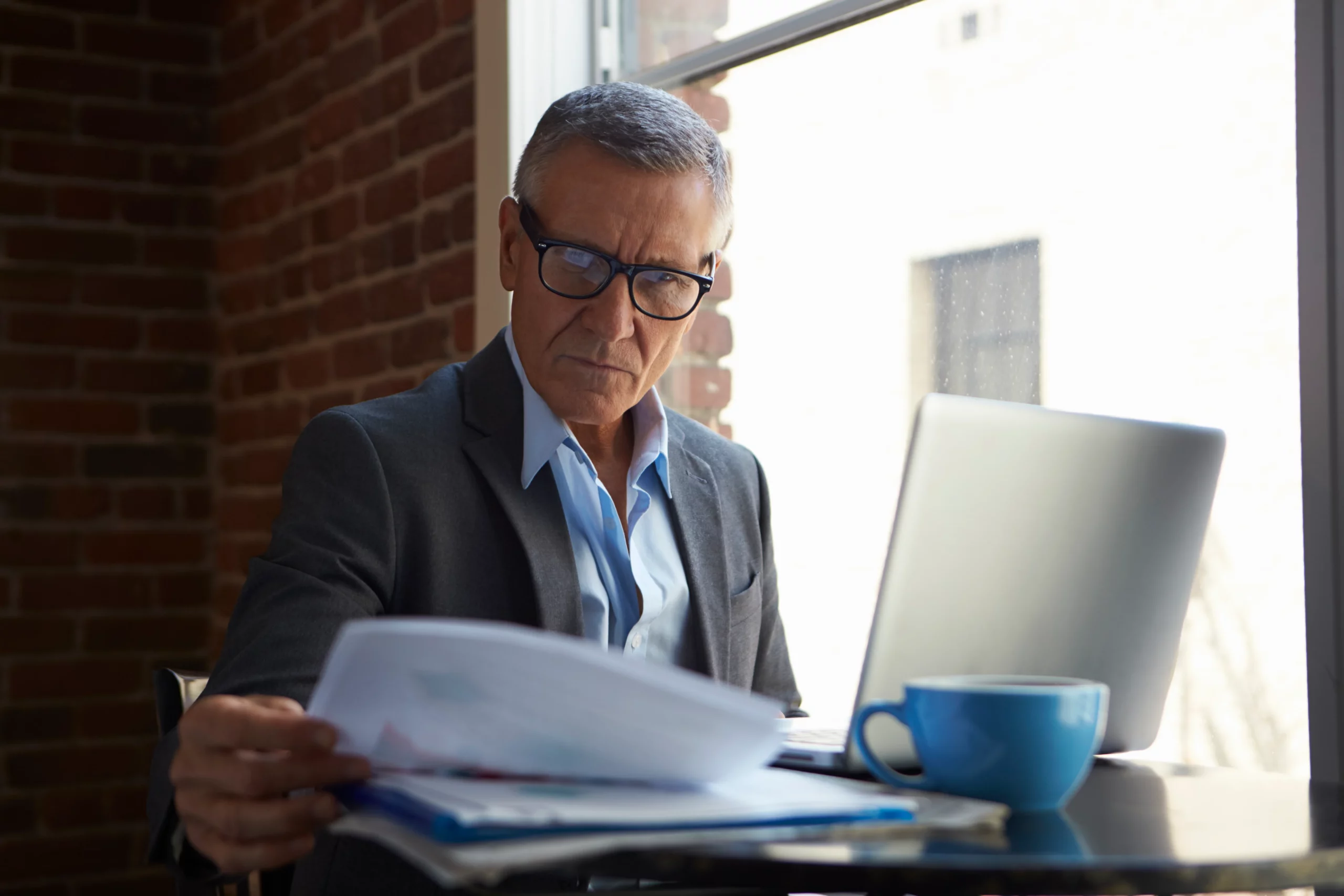 Older man in suit reviewing papers at a table by a window with laptop and mug, illustrating careful review of trustee personal liability in California trust law.