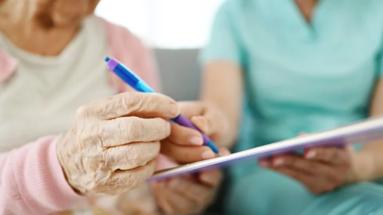 Elderly woman’s hands holding a pen to sign a document with caregiver in scrubs assisting, representing concerns of undue influence on wills and estate planning in California